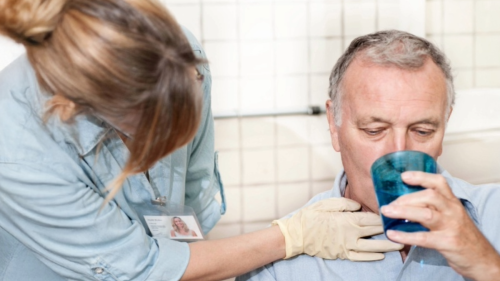 A man drinks from a cup of water while a healthcare professional examines his neck