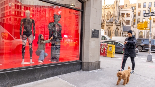 a students adjusts a mannequin in a window as a dog walks by