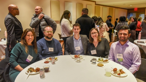 Five people sit around a table. Behind them, people stand and chat.