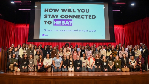 A large group of attendees gathers on a stage beneath a screen that says "How will you stay connected to HESA?"