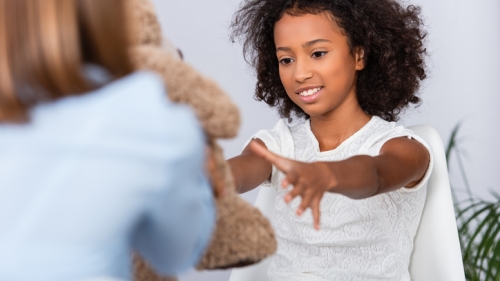 Image captures a child sitting in a chair. Across from her is an adult holding a brown stuffed animal. The female sits across from the adultl, reaching across with her arms fully extended to receive the stuffed animal from the adult. The adult wears a blue colored blouse and has blond hair. Only the child's face is visbile to the camera lens. The photographer only captures the back of the adulting holding the stuffed animal.