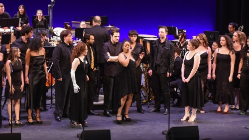 A female singer sings into a microphone, as other singers watch on.