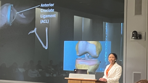 Dannyelle Long stands at a podium; behind her, a screen shows an image of a knee with the label Anterior Cruciate Ligament (ACL)