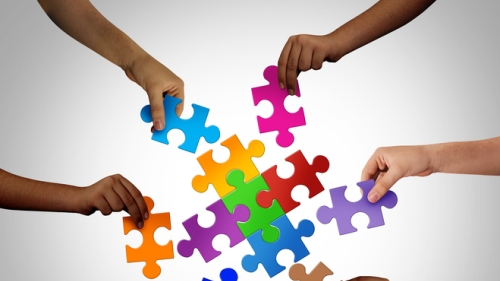 Image captures hands and arms of six children putting puzzle pieces together. Each of the 6 puzzles pieces are of different colors. The coming together of the these individuals from diffferent cultureal backgrounds signiefies diversity, quity, and inclusion.