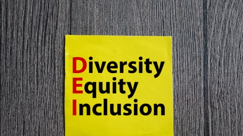 Image captures a square shaped image with text. The graphic features a large yellow post set against a Black colored wall. The Blck wood panel wall has a yellow post with predominately black text on it. The text on the post reads "Diversity, Equity, and Inclusion. The first letter of each of those 3 words are both captilized and appear in read colored text. The 3 words, Diversity, Equity, and Inclusion are verically stacked so that the red letters (from top to bottom read), DEI.