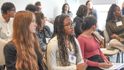 This snapshot captures attendees of NYU Metro Center's 2025 Equity Conference. The attendees make up the audience for breakout sessions.. Th photo captures more thn 12 individual in the breakout session. The image itself is centered around3 figures un the frount row. The figure on the lleft wears a black blazer. She also has long red hair. The figure inthe middle wears a white toop and sports lon black braids. This inidivual also has items in her lap to take notes during the session. Lastly, the indidual t 