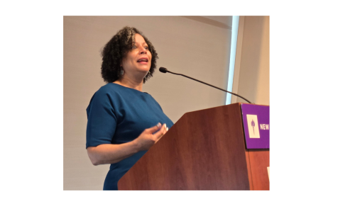 The snapshot captures Dr. Fabienne Doucet addressing NYU Metro Center's 2025 Equity Now Conference.. Dr. Doucet speaks to the conference attendees from the dias. She wears a teal colored dress, and sports her naturally curly hair. She speaks from a wooden lecturn with the NYU logo on the front. The thin microphone is drawn back to capture each of her words.