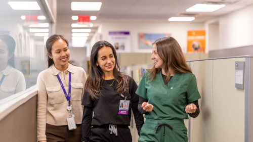 Three students, two in scrubs, walking and chatting in a hallway.