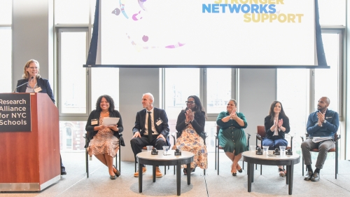 One person stands at a podium. Six panel members sit and applaud. Behind them, a screen reads, “Weaving Stronger Networks of Support.”
