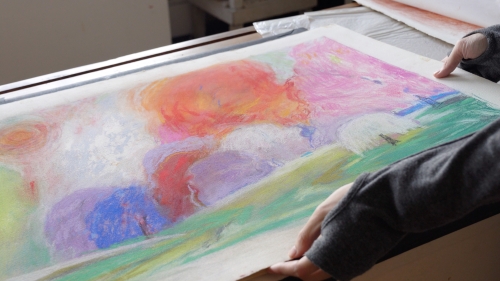 Two hands hold a colorful pastel drawing of a landscape