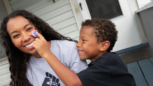 Image captures a mother and son at play in front of their home. Both the mother and son sit out the steps of their home, as they sit in mid embrace. The son wearing a black shirt, reaches towards his mother's face with his index finger. The mother wearing a gray colored shirt, with a Black-Power icon t-shirt on, smiles as her son gleely attempts to place an "I voted" sticker on her nose.