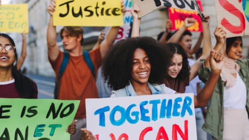 Image captures youth protesting in the street. This group of protestors are in the street holding signs with text that express their calls for change. While the photo itself is a wide shot that captures a total of ten protestors, the picture actually centers around black female protestor. She is in the middle of the frame and holds a sign which reads, "Together we can change". .