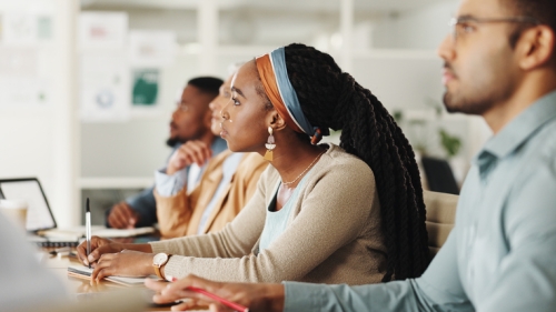 Image captures a Black Woman educator seated at a long row table with fellow instructors. The Black Woman looks intently focused on the lesson going on at the front of the classroom. She wears a khaki colored sweater over a white shirt. She is wearing her hair in braids and wears a blue and brown colored headband. The educator seated to her left wears a light blue denim shirt and eye-glasses.