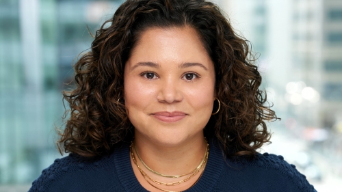 A latina woman in a blue sweater smiles at the camera. She has brown shoulder length hair. The background shows a blurred city street.