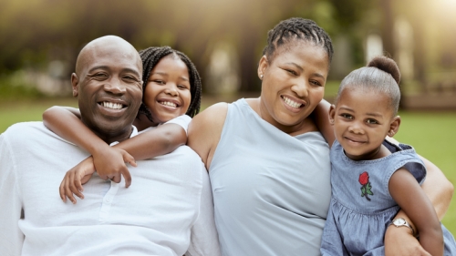 Image captures a family gathered together in a park. The photo is centered around a mother and father sitting in a green space with their two young daughters. To the right of the photo, the father sits facing the camera, as one of his daughter hugs him from behind.. He wears a white t-shirt, and she smiles over his left shoulder. On the right, the mother sits adjacent to her young daugher.. The mother waes an aqua colored top, while the daughter wears a blue top..