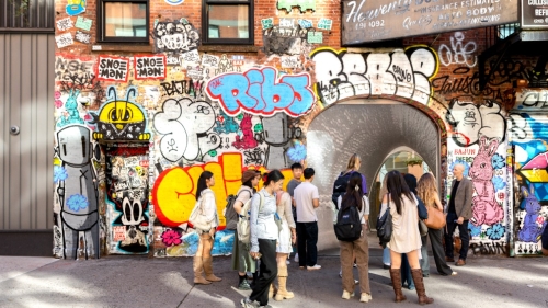 Students stand on a sidewalk next to a graffiti-covered wall