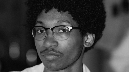 In this black and white portrait image, the artist is wearing glasses, a white shirt, and has a mustache. 