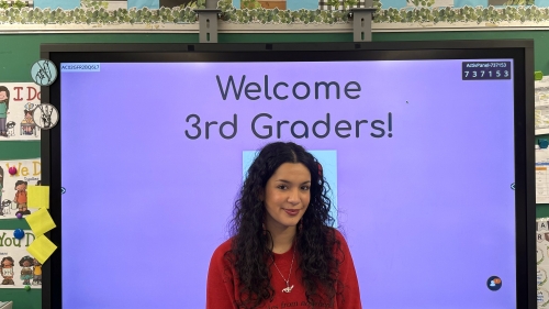 Gabriela stands in front of her classroom. The screen behind her reads, "Welcome 3rd Graders!"