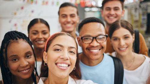 Image captures seven multiracial high-school aged youth, gathered together to take selfie. The youth at the center of the image has a white floral top. To her left, a fellow student wears a blue t-shirt and back. While the student to his left wears white scoop neck t-shirt. The other four individuals in the photo are seen only from the neck up. The right of the youth in the center, is a with black briads down to. her shoulder. 
