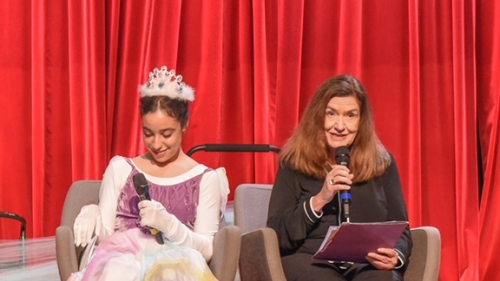 Two women on stage with microphones. The woman on the left is wearing a crown and princess costume. 