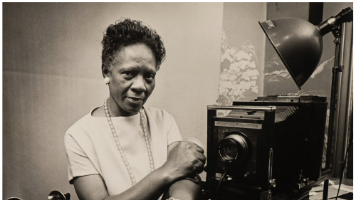 An African American older woman is show next to an old fashioned B&W studio camera.
