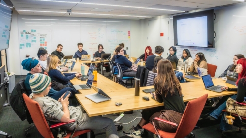 Students sit around a conference table on their laptops or phones while they listen to Professor Safinah Ali