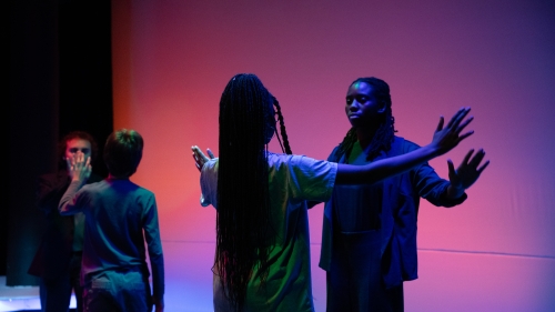 Four actors in pink tinted room performing in pairs facing one another