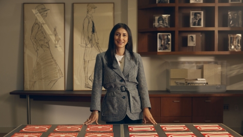 A woman with black hair smiles facing the camera. She is standing behind a table with printouts of red graphics.