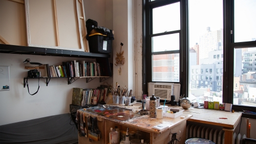 An artist studio featuring a desk center frame with paint brushes and supplies on it, a book case on the left, and large windows looking out into the city on the right.