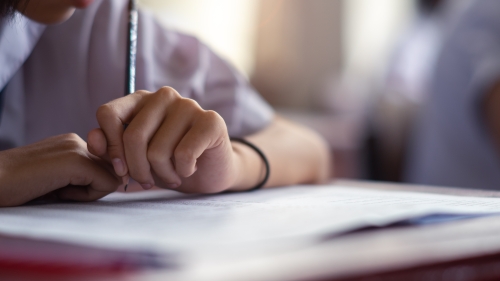 A hand holding a pencil hovers above at paper at a desk in a classroom.