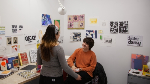 Two students talking in a BFA studio, colorful artwork is pinned on the wall behind them