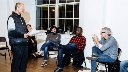 A performer stands, holding a piece of paper, getting feedback from an instructor while a small, seated group observes