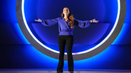 Student standing in front of large blue lit up circle with arms spread and smiling