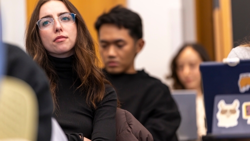 Student in foreground looking up to front of classroom sitting at desk and two students behind her also at desks. 