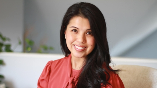 Headshot of Aimee Hilado smiling at the camera, wearing a coral blouse with a tie detail, with long dark hair and a softly lit indoor background with plants.