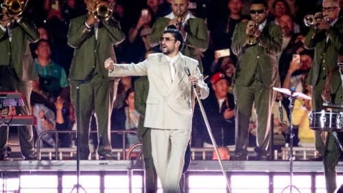Bad Bunny on stage in front of a live band, performs in a tan suit