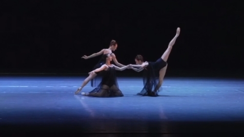Three dancers on a dark stage perform in black leotards and sheer skirts
