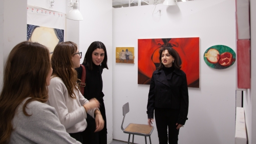 A group of students stand in a studio with artwork installed on the wall. One student wearing all black answers questions while standing in front of a large red painting.