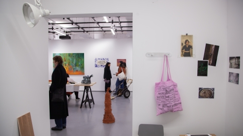 Artist studio with a pink tote bag hanging on a bag rack and printed photos pinned to the wall studio. Outside of the studio, viewers look at other student work including a tall brown ceramic piece on the ground.