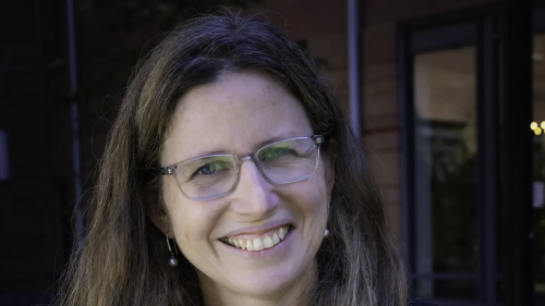 Headshot of Milagros Nores smiling at the camera, wearing glasses, a dark blazer, and a statement necklace, with long light brown hair and an outdoor building background.