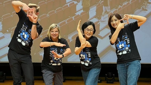 4 faculty wearing matching shirts stand on a stage and make hand and arm gestures, behidn them is a projected image of empty stadium seating