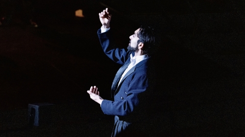 Solo performer on black stage. Only light is him in the bright spotlight. Very dramatic.
