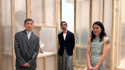 Three people smiling in the camera. They are standing indoors in between structures made with plywood.