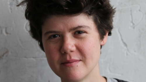 Headshot image of Cate standing in front of a white textured wall, wearing a black long sleeve sweater over a gray shirt.