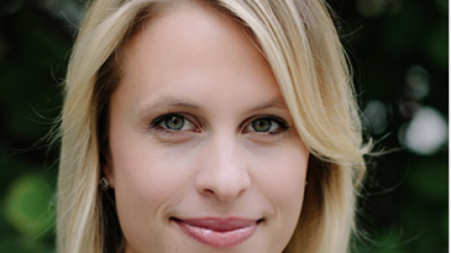 Headshot image of Elle, she has blond hair and is wearing a navy coat. She is smiling center frame and standing in front of greenery which is out of focus.