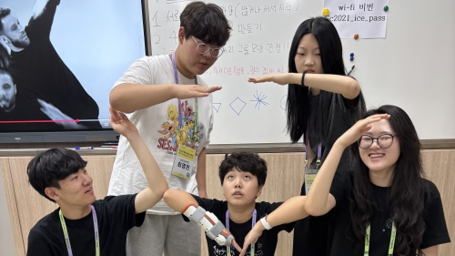 5 students stand and kneel in a classroom to make a triangle shape using their arms