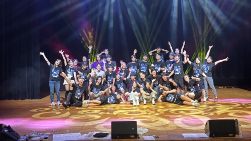 A large group of people pose for the picture on a stage, standing in a group with arms out under bright stage lights
