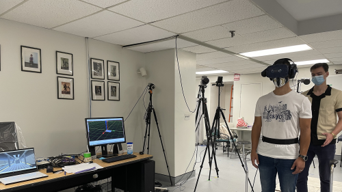Participant stands on balance board while wearing VR headset, while one research assistant stands behind them and nearby computers run the experiment