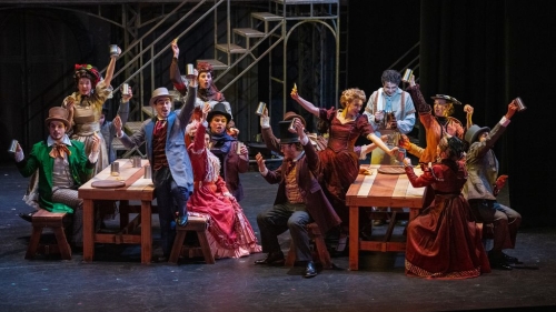 Actors on stage sitting at two long tables singing and toasting each other