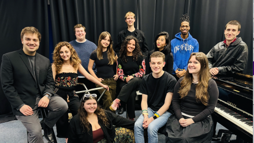 Group photo of the Village Voices ensemble posed around a piano